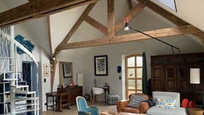 Un salón espacioso con techo alto y vigas de madera a la vista, un área de descanso acogedora con un sofá y sillones, una mesa de centro de madera y una estantería con objetos decorativos.