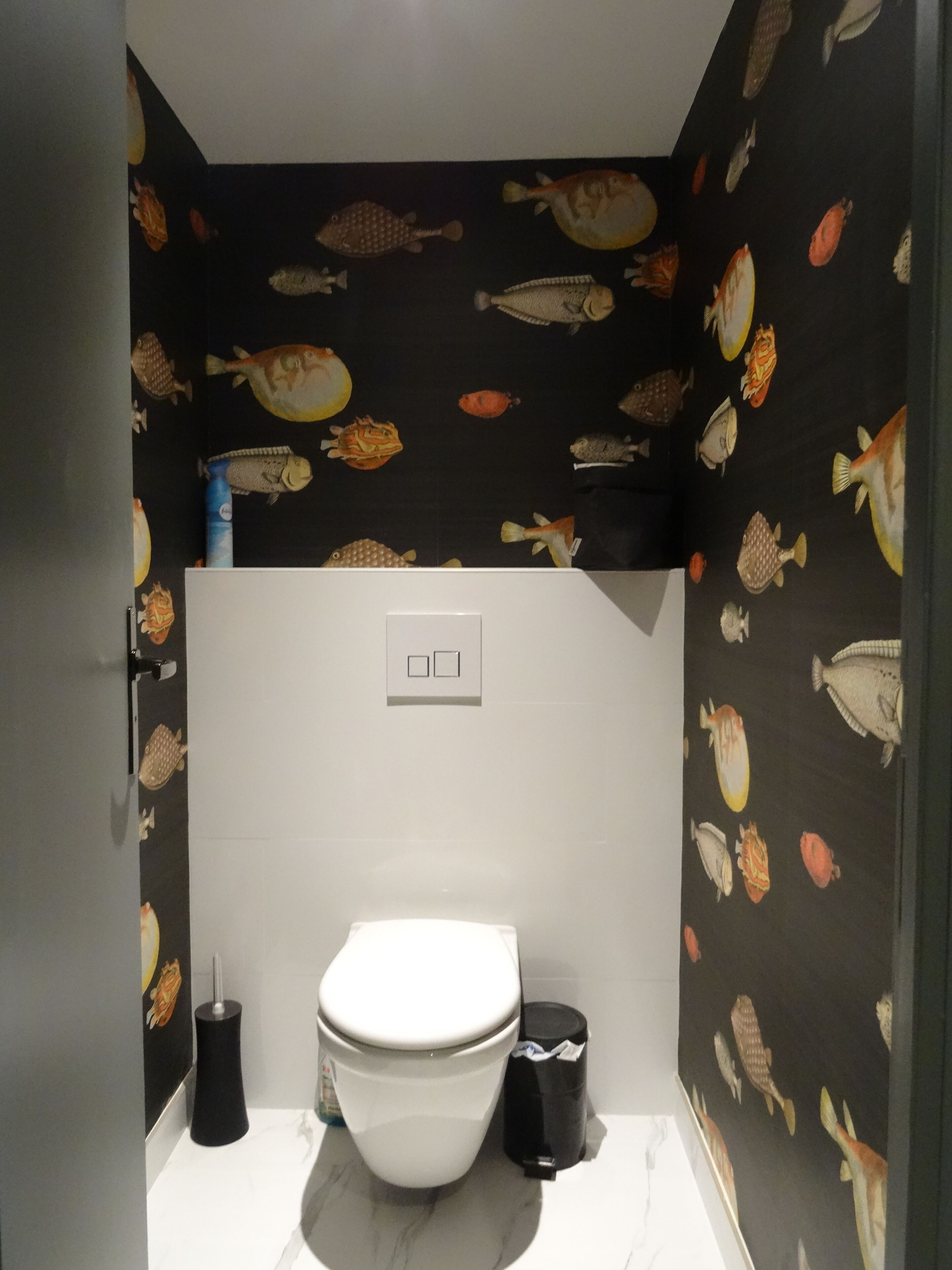 A small bathroom with a toilet, a trash bin, and a wall with fish patterns.