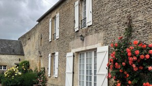 A stone building with white shutters, a gravel driveway, and flowering plants. - Pretty stone house, 3 bedrooms, 2 bathrooms with a large enclosed garden (Longues-sur-Mer)