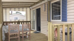 Un espace extérieur abrité, doté d’une terrasse en bois, d’une table avec des chaises et offrant une vue sur une maison à travers une fenêtre.