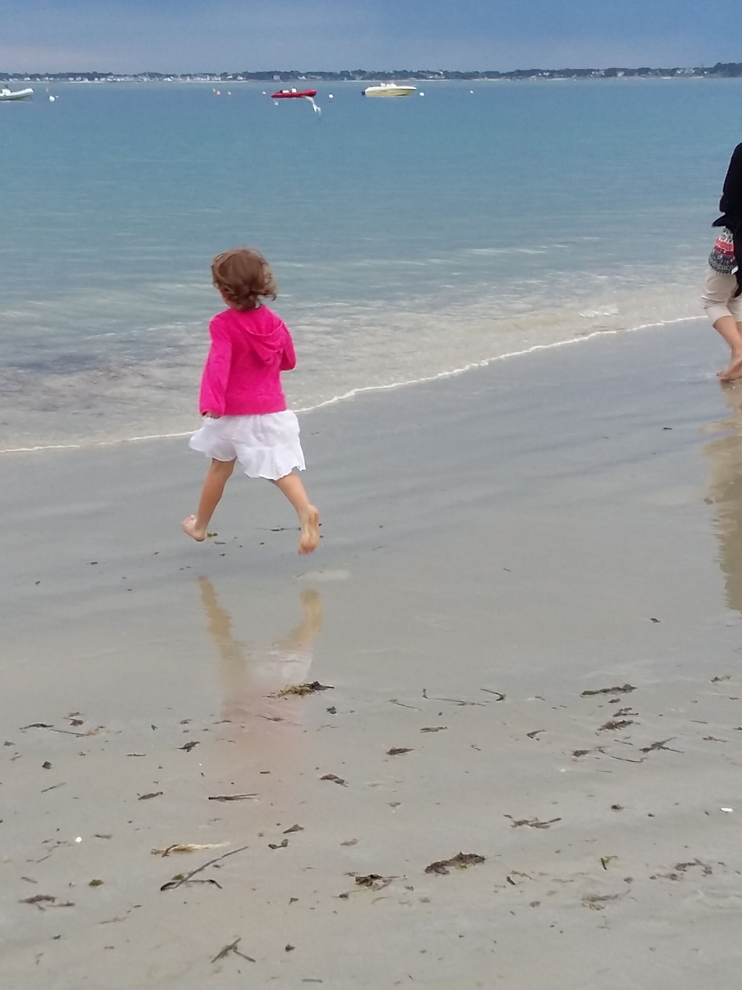 A beach scene with a child in a pink top and white skirt walking along the shore.