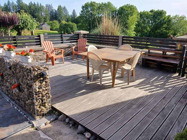 Una terraza de madera con mobiliario exterior, un muro de piedra y vistas a una casa y árboles.