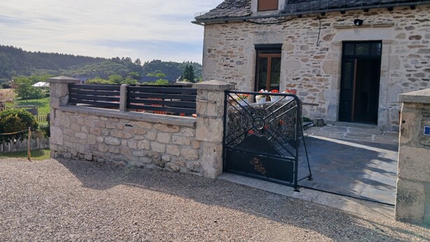 A stone building with a black metal gate, a window with a flower box, and a view of a forested area.