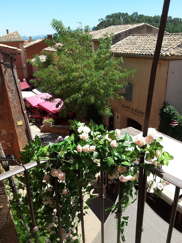 A balcony with a climbing rose plant, a building with a sign reading "BLANC & BLANC Project Management," and a red umbrella in the background. - "LA DAME AU BALCON" on the village square (Roussillon)