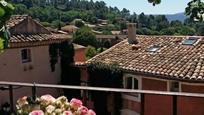 A balcony with a vase of pink flowers, overlooking a town with traditional buildings and a clear blue sky. - "LA DAME AU BALCON" on the village square (Roussillon)