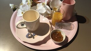 Un plateau avec une tasse, un verre de jus d’orange, un petit bol de confiture, un panier de viennoiseries et des fleurs blanches.