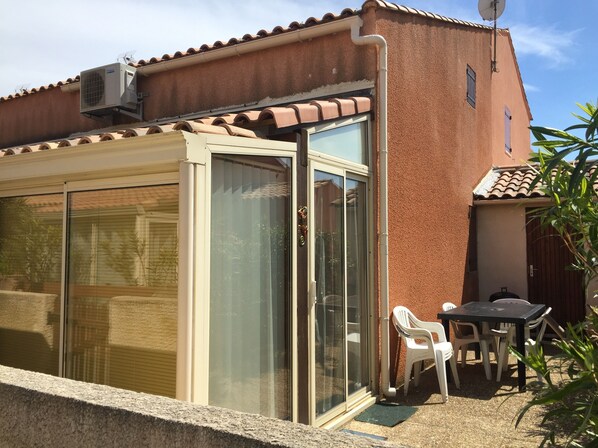 Un edificio con tejado de tejas, puerta corrediza de vidrio y un pequeño patio con sillas de plástico.