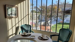 A dining area with a table set for two, a framed picture on the wall, and a view of a garden through large windows.