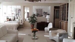 A spacious living room with a high ceiling, a chandelier, white furniture, a coffee table with a vase of flowers, and a view of the kitchen through a glass door.