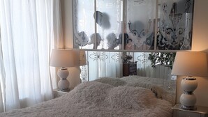A bedroom with a fluffy white bedspread, two bedside lamps, a large window with decorative curtains, and a wall with a floral pattern.
