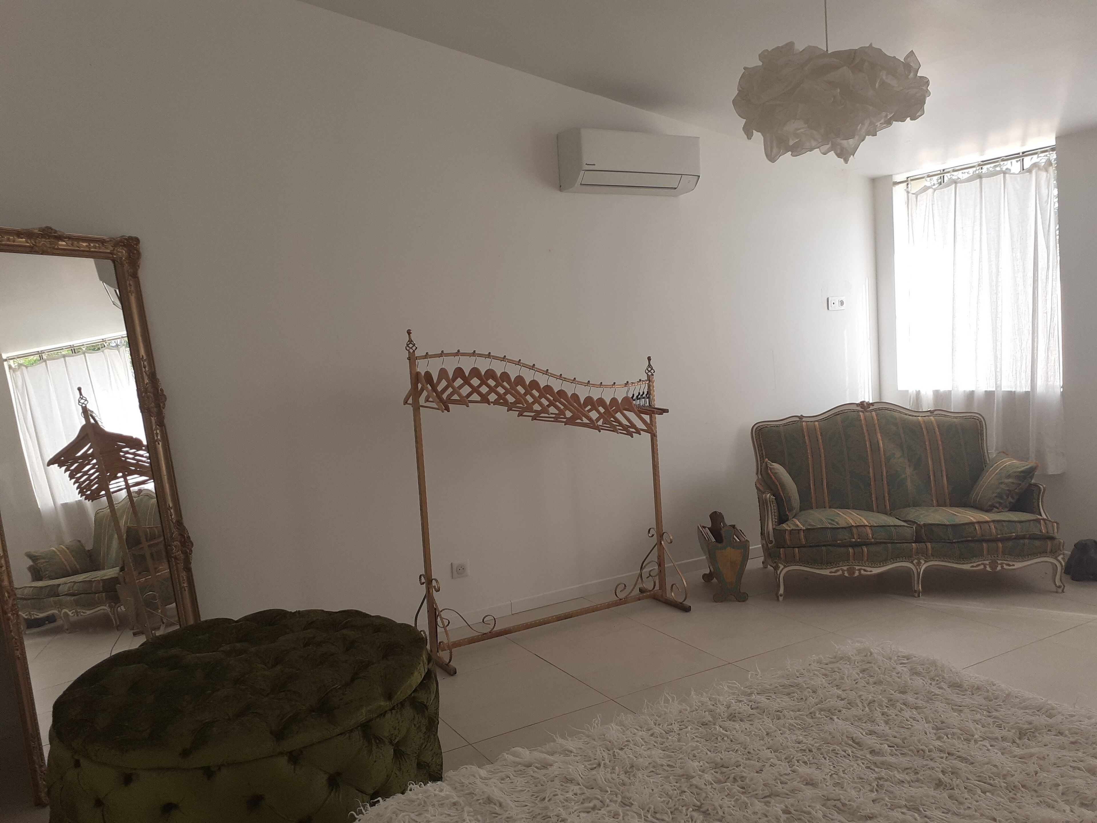 Quarto com um banquinho estofado verde, um sofá estampado, um varal de madeira, um ar condicionado branco e um candelabro.