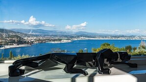 A view of a coastal city with a clear blue sea, mountains in the distance, and a modern building with a unique architectural design. - ★ House with Panoramic Sea View ★ (Théoule-sur-Mer)