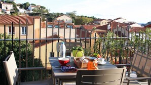 A balcony with a table set for a meal, surrounded by a metal railing, overlooking a residential area with terracotta roofs.