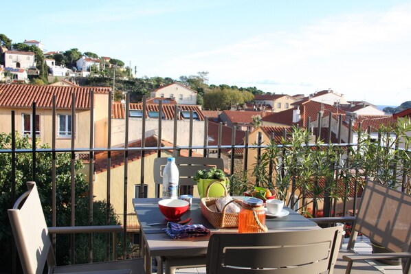 Un balcón con una mesa lista para comer, rodeado por una barandilla de metal, con vistas a una zona residencial con tejados de teja.