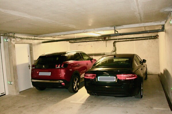 Dos coches estacionados en un garaje subterráneo con paredes de hormigón y una puerta blanca.
