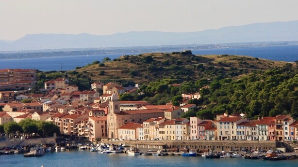 A coastal town with historic buildings, a marina, and a hill with dense greenery in the background.