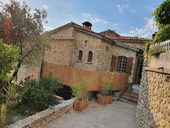 A stone building with a tiled roof, wooden shutters, and a small garden area with potted plants.