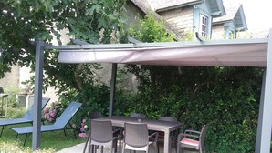 Een buitenpatio met een overdekte tafel en stoelen, omgeven door groen en een huis op de achtergrond.