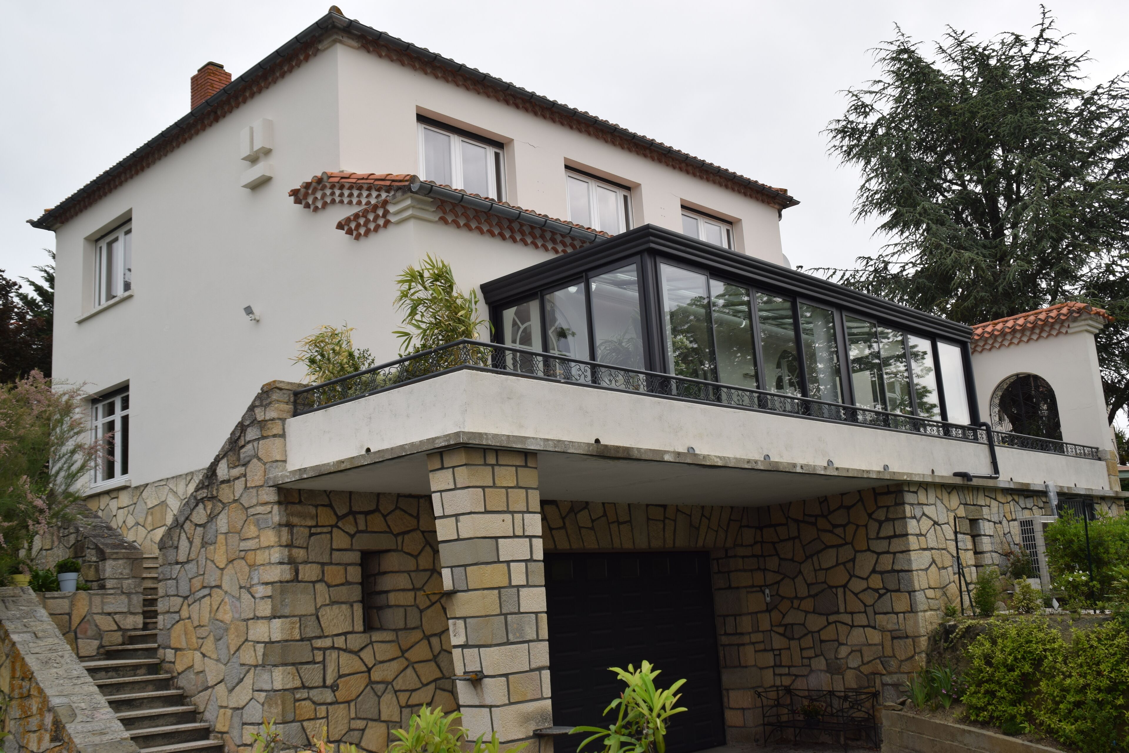 A two-story house with a stone facade, a glass conservatory, and a staircase leading to the entrance.