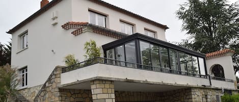A two-story house with a stone facade, a glass conservatory, and a staircase leading to the entrance.
