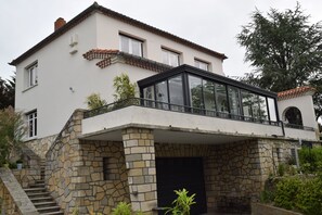 Uma casa de dois andares com fachada de pedra, uma estufa de vidro e uma escadaria que leva à entrada.