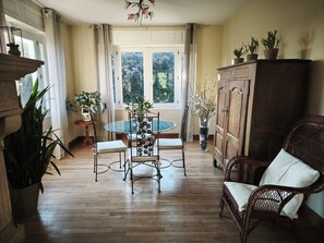 Ein Zimmer mit einem runden Glastisch, zwei Stühlen, einem Korbssessel, einem Schrank und Topfpflanzen.