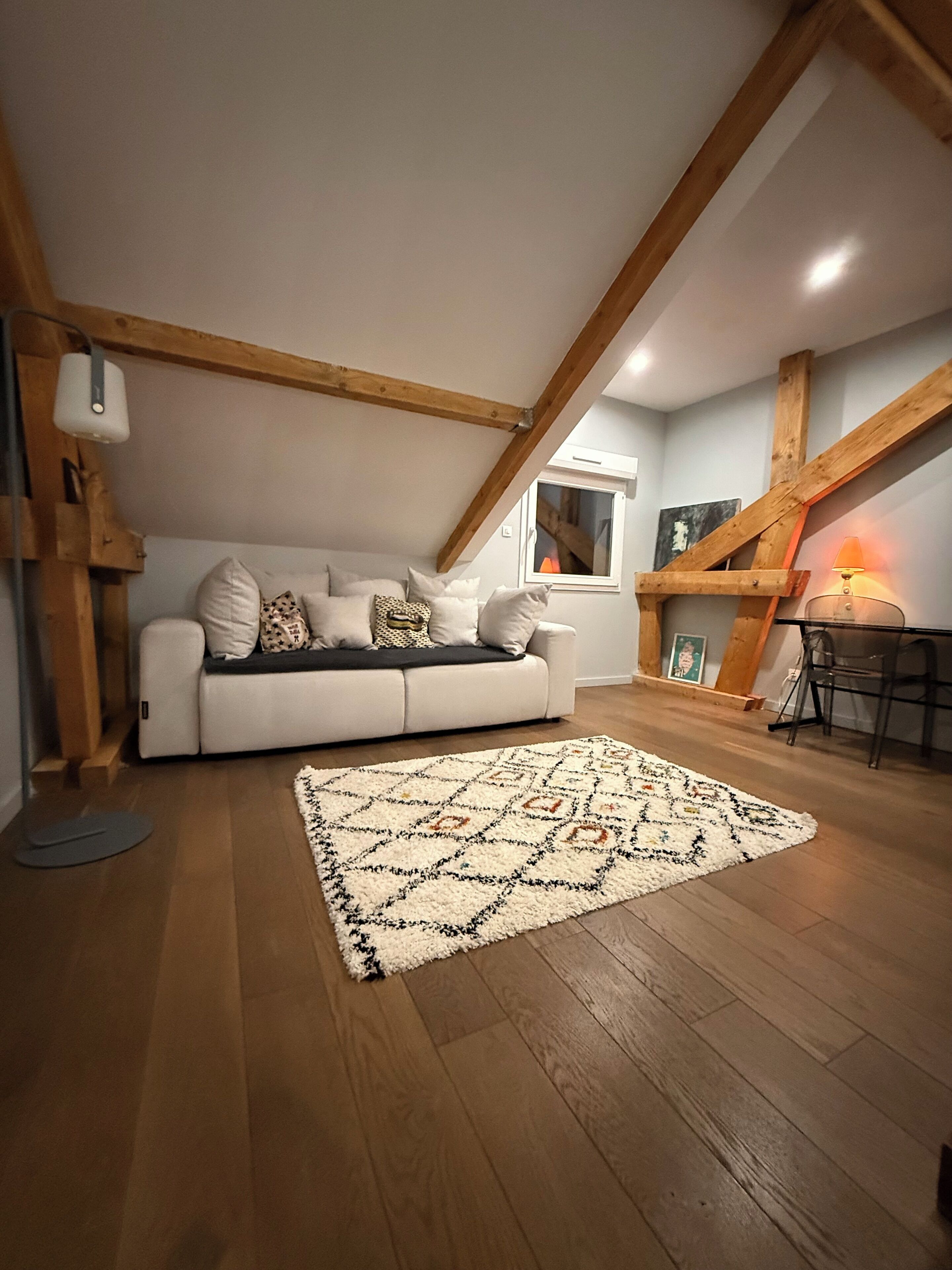 A cozy loft bedroom with a white sofa, patterned rug, wooden beams, and a small table with a lamp.