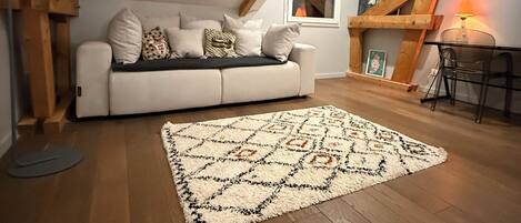 A cozy loft bedroom with a white sofa, patterned rug, wooden beams, and a small table with a lamp.