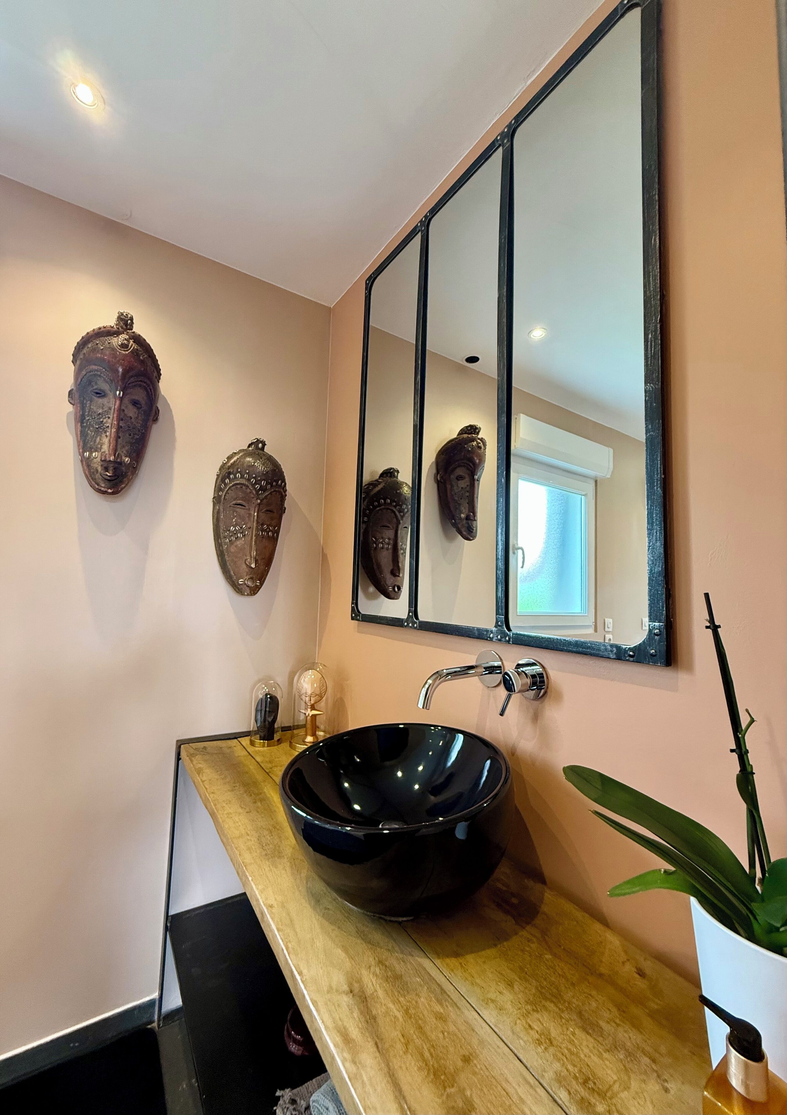 Un baño con un lavabo tipo cuenca negro, un mueble de tocador de madera, un espejo grande y dos máscaras decorativas en la pared.