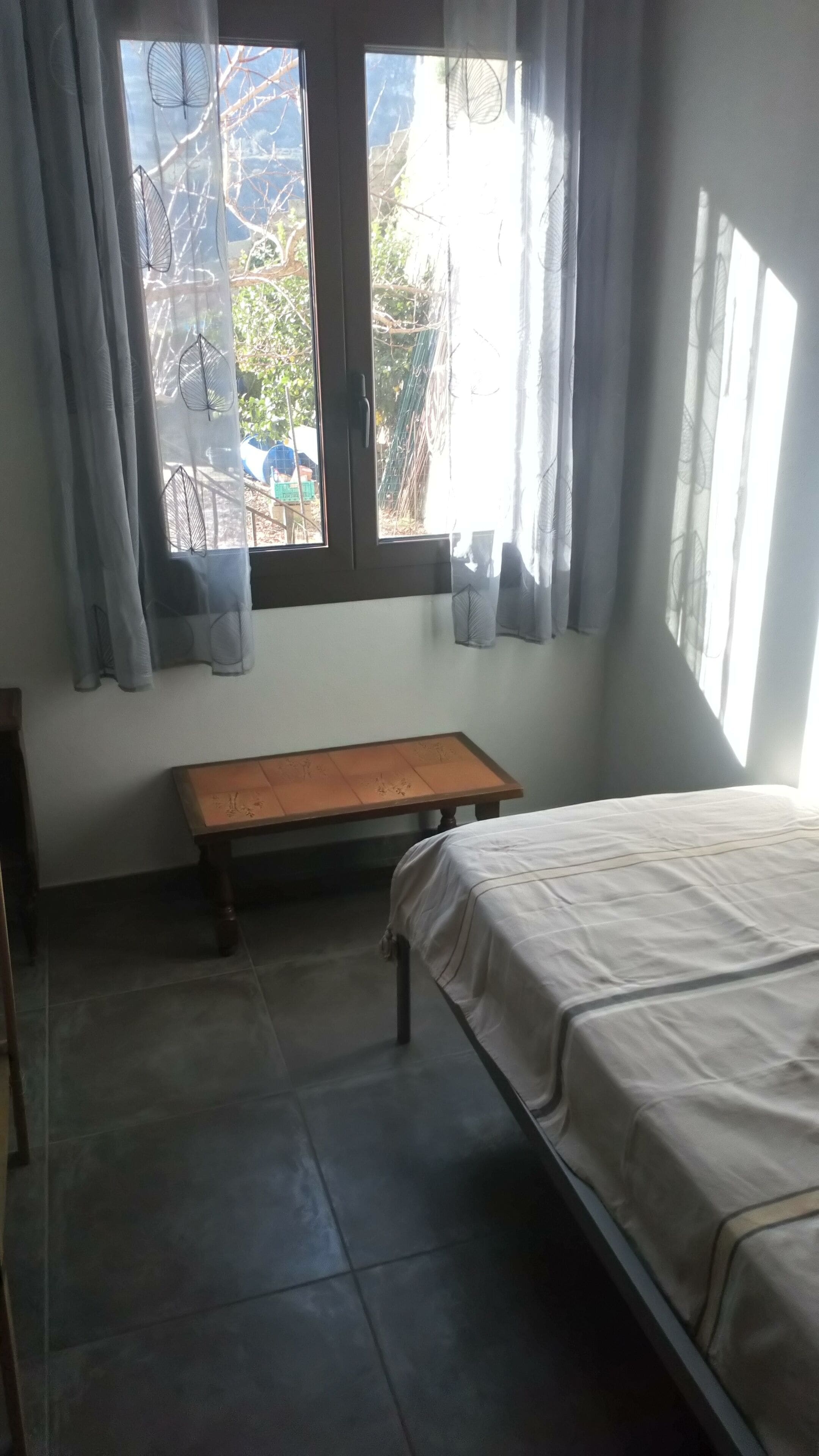 A bedroom with a bed, a wooden bench, a window with curtains, and a view of trees and a fence outside.