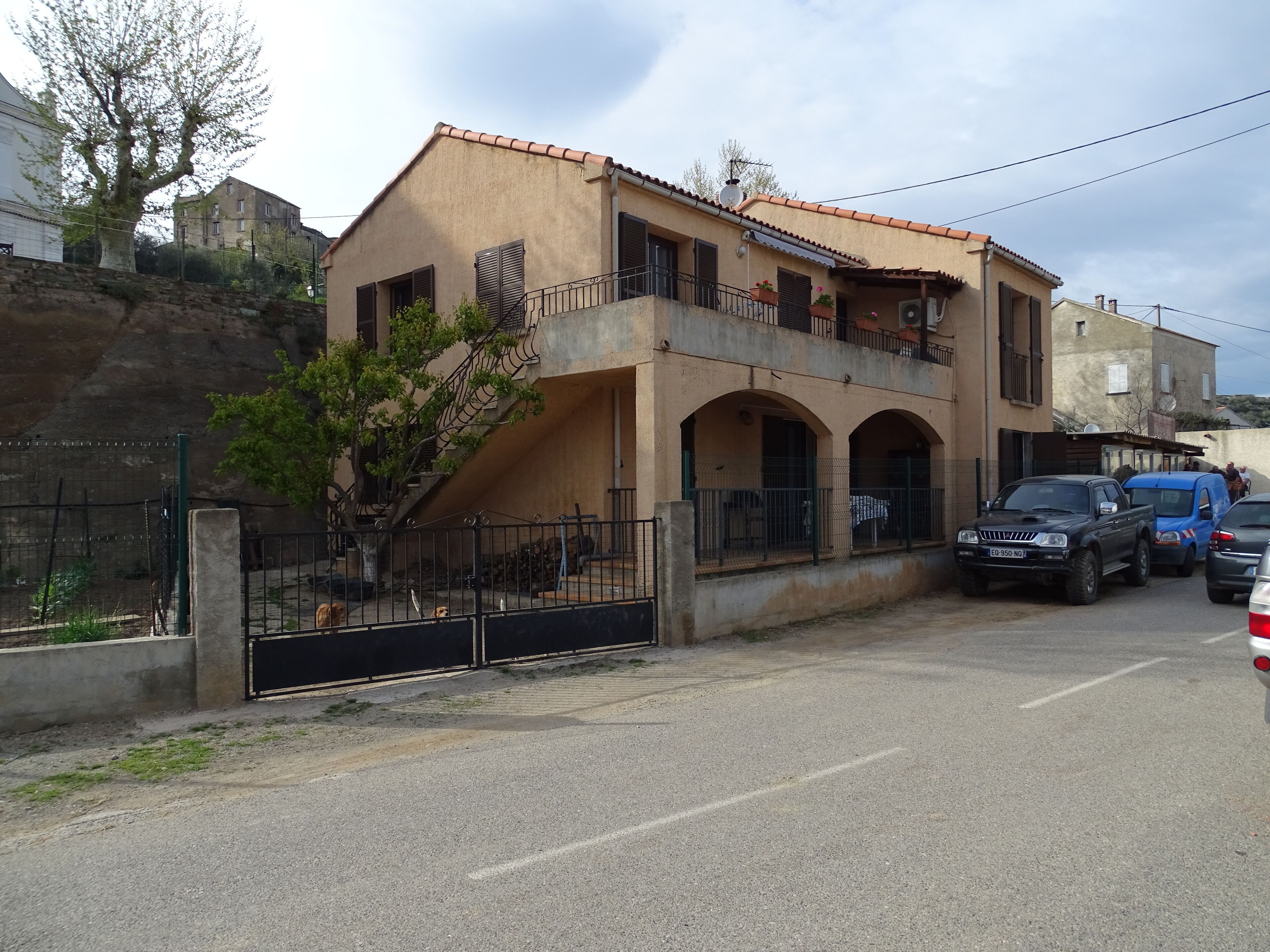 Un immeuble à deux étages doté d’un balcon, d’une entrée sécurisée par un portail et de voitures garées.