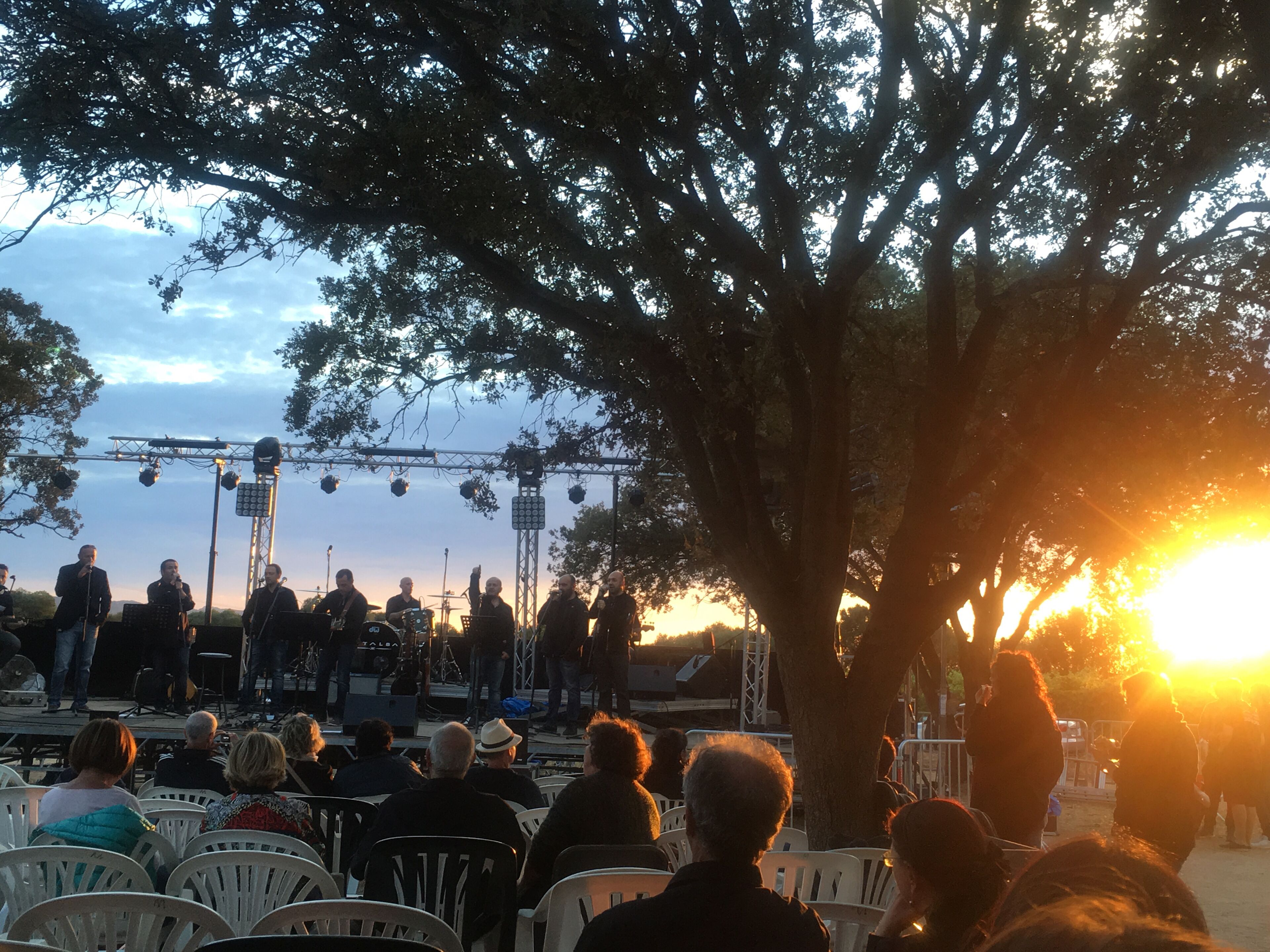 Um palco com músicos apresentando-se, rodeado por árvores e uma plateia sentada em cadeiras brancas.