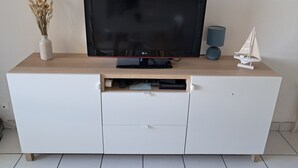 A flat-screen TV on a wooden TV stand with a drawer, a vase of dried plants, and a small sailboat model. - F2 250 m from the beach of Chay Royan (Royan)