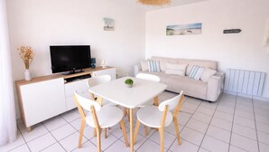 A modern living room with a white sofa, a dining table with chairs, a TV on a wooden cabinet, and a hanging light fixture. - F2 250 m from the beach of Chay Royan (Royan)