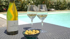 A bottle of Domaine Perdrix-Lasouque wine, two glasses of white wine, and a bowl of olives on a table by a pool.