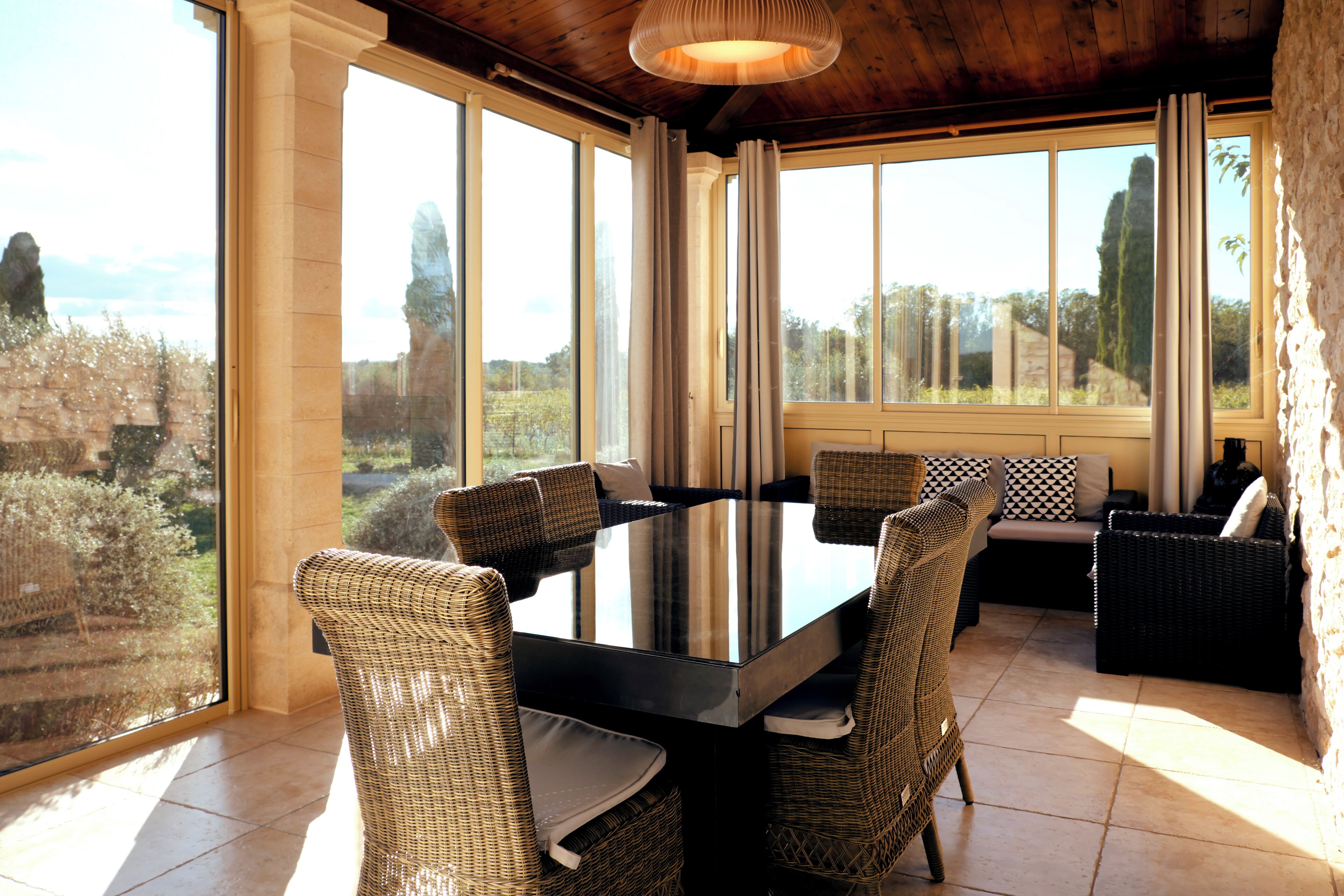 A dining area with a glass table surrounded by wicker chairs, large windows, and a view of the outdoors.