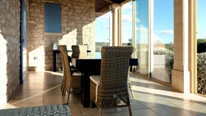 A sunlit patio with stone walls, wicker furniture, and large windows offering a view of the landscape.