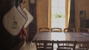 A dining area with a wooden table and chairs, a hanging decorative item, a clock, and a window with curtains. - Maison De La Residence Celeste (Ogenne-Camptort)