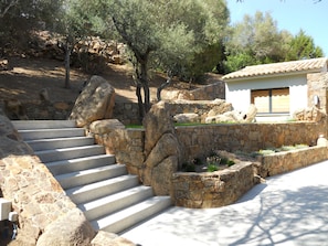 A stone staircase leading up to a building with a tiled roof, surrounded by large rocks and olive trees.
