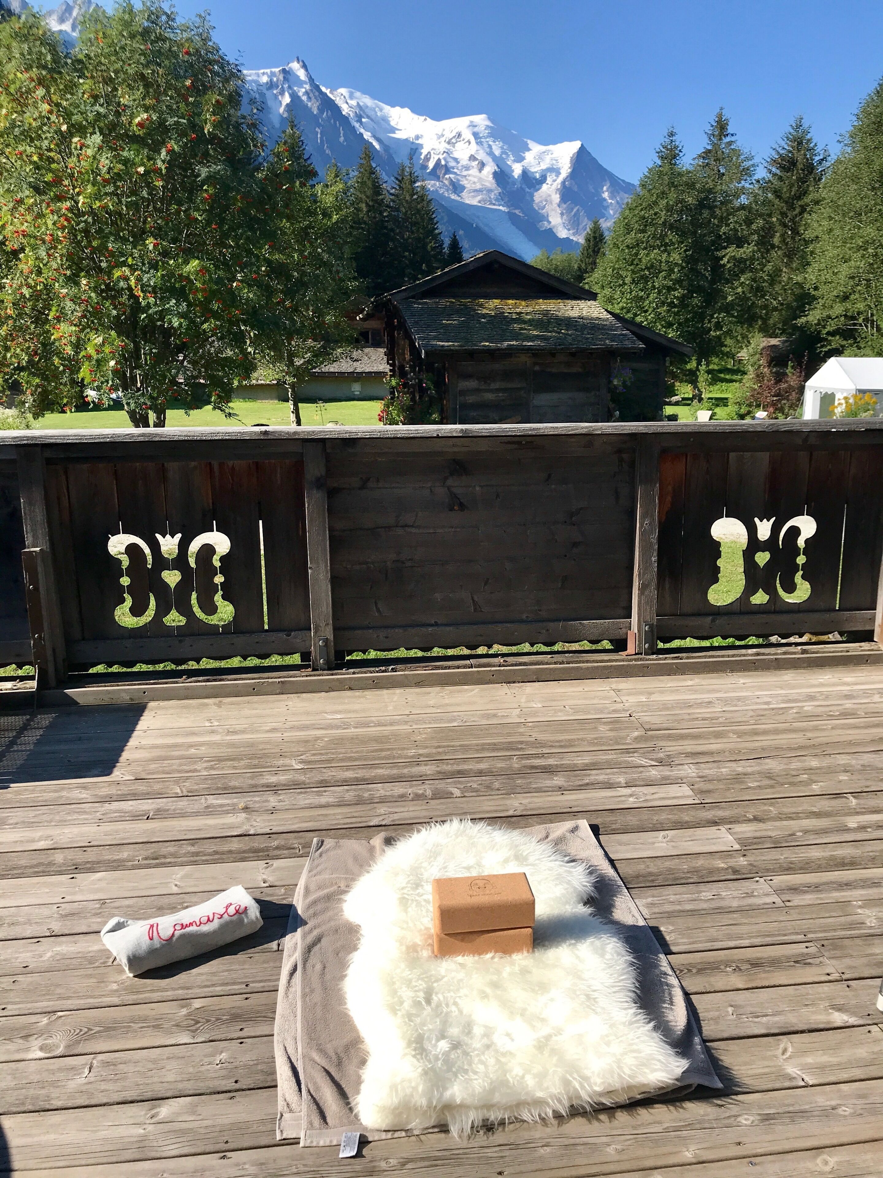 Una terraza de madera con un almohadón blanco y esponjoso y una caja marrón, una valla de madera con grafiti, y una montaña al fondo.