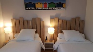 A hotel room with two beds, wooden headboards, white bedding, and a painting of colorful beach huts above the beds.