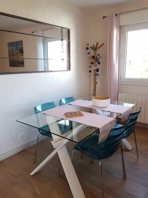 A modern dining area with a glass table and teal chairs, a white bowl on the table, and a decorative tree on the wall. - Cap Meyran - T3 Cosy with Pool (Gujan-Mestras)