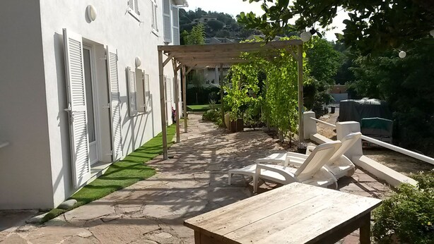 Eine Terrasse mit einem gepflasterten Weg, weißen Fensterläden, einer Pergola und Gartenmöbeln.