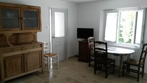 Ein Zimmer mit einem Holzkasten, einem kleinen Hocker, einem Tisch mit Stühlen, einem Fernseher auf einem Schrank und einem Fenster mit Blick auf Grünflächen.
