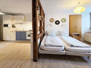 A compact room with a bed, a wooden headboard, a small bedside table, a wall-mounted lamp, and a kitchen area in the background. - Rempart - Large studio in the centre of the village (Eguisheim)