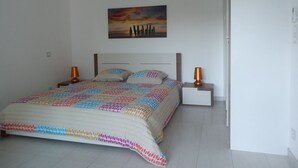 Una camera da letto con un letto, due comodini, un tavolino e un condizionatore d'aria a parete, oltre a un quadro raffigurante un tramonto.