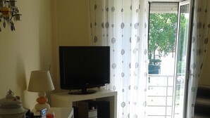 Sala de estar con televisor de pantalla plana sobre un mueble bajo, una lámpara y una mesita con adornos. Hay un ventanal corredero que da a un espacio exterior.