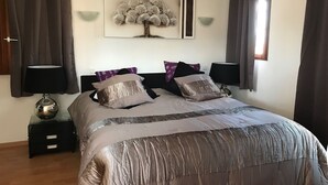 Une chambre à coucher avec un lit, deux tables de chevet avec des lampes, une œuvre d’art représentant un arbre accrochée au mur, et une fenêtre avec des rideaux.