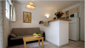 Un salon comprenant un canapé, une petite table en bois avec un saladier de fruits, un luminaire fixé au mur, un tableau, et un coin cuisine avec un four à micro-ondes et un réfrigérateur.