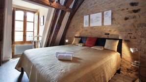 Un dormitorio con pared de piedra, vigas de madera, una cama con colcha color beige y una mesita de noche con lámpara.